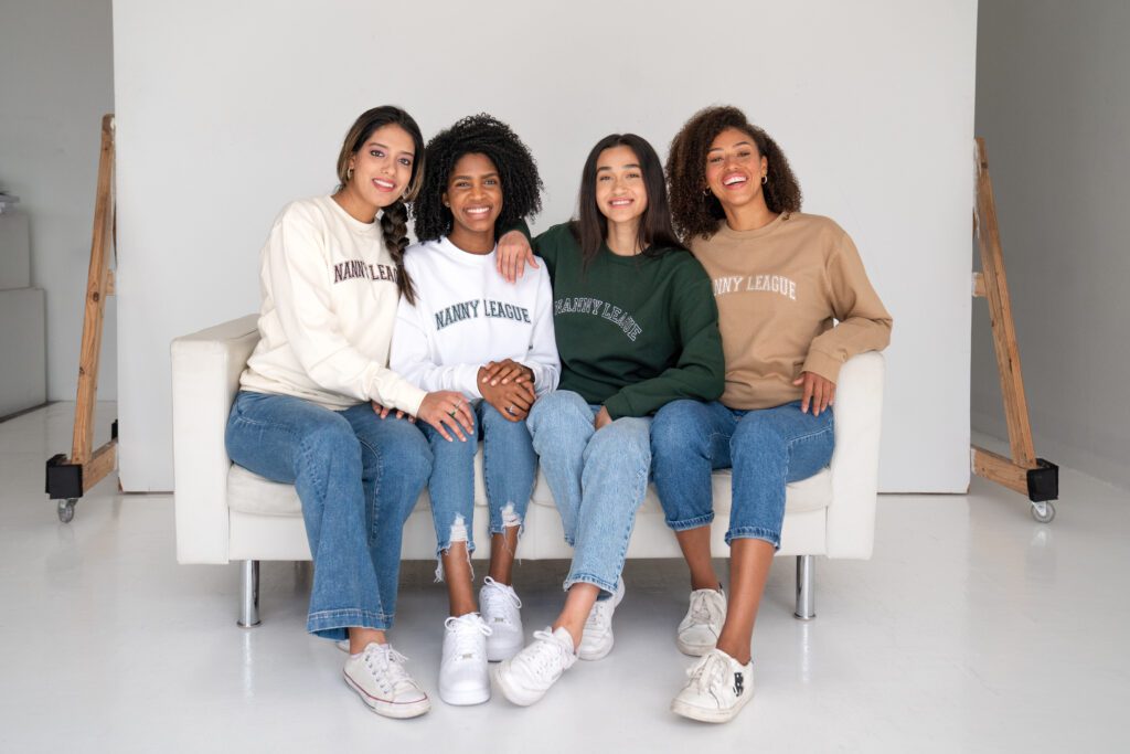 Four nannies sitting on a couch smiling at camera while wearing The Nanny League sweatshirts